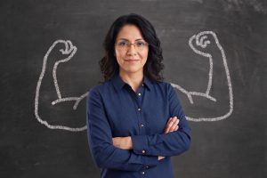 Professional woman with glasses and blue shirt stands confidently with arms crossed in front of a chalkboard drawing of flexed arms.